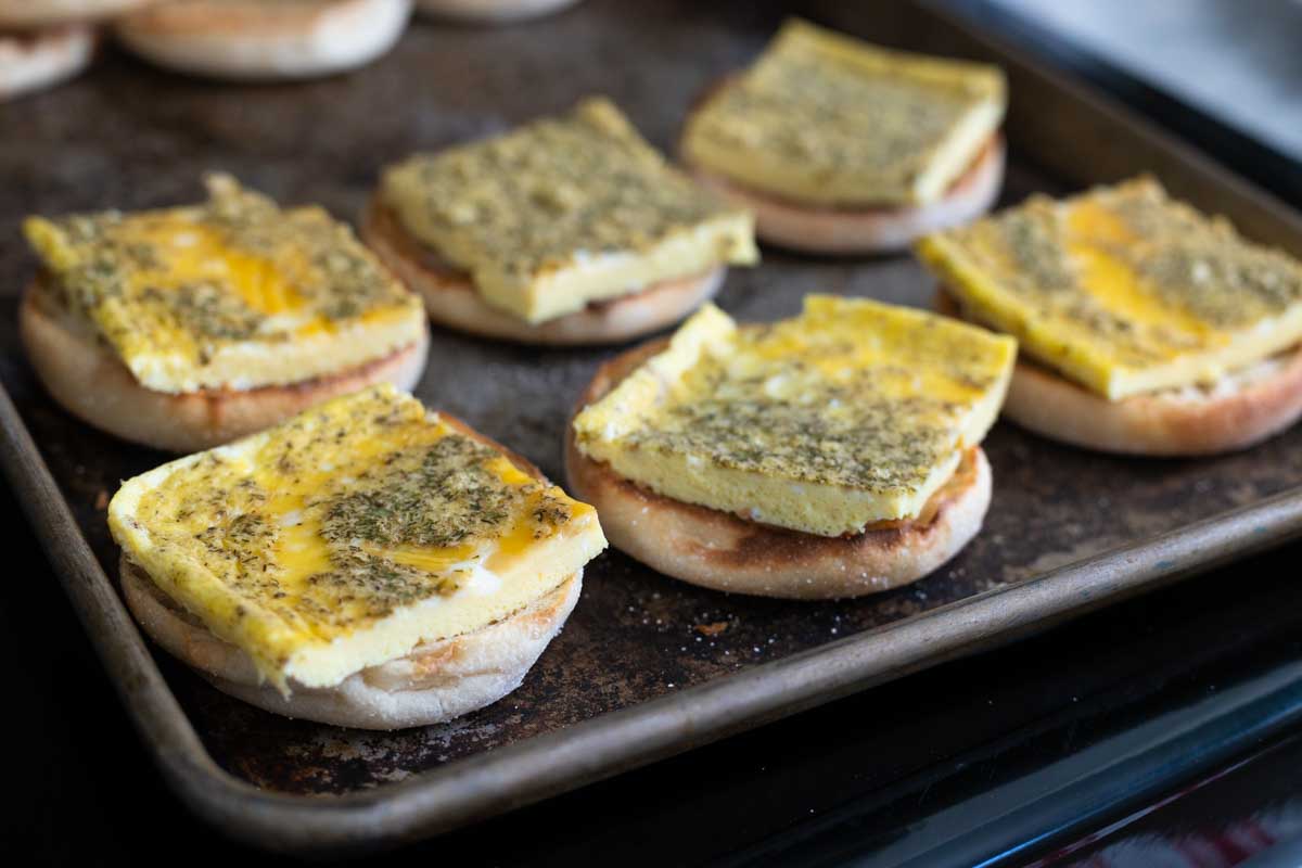 The egg patties have been sliced into squares and added to broiled english muffins on a baking sheet.