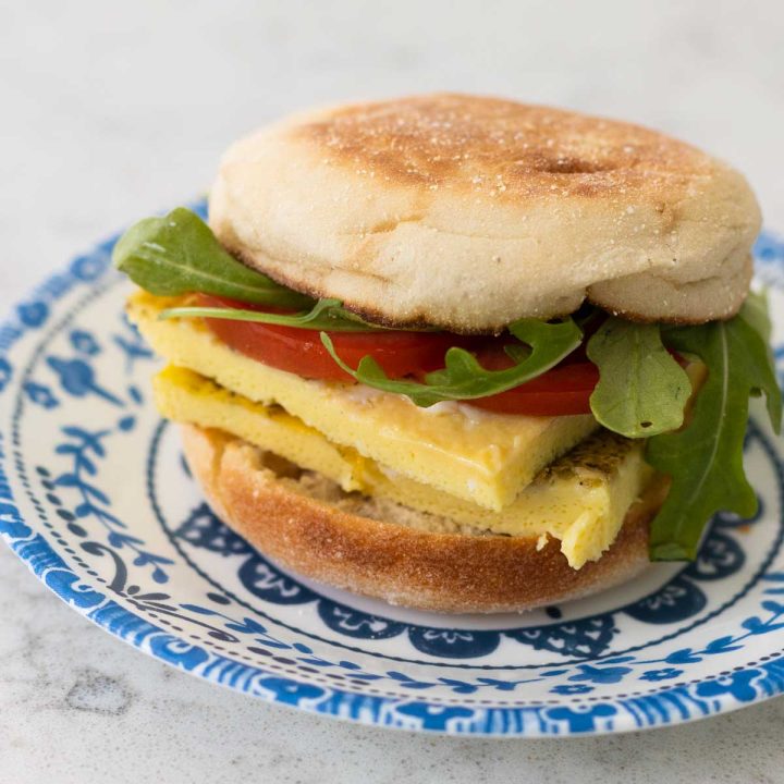 A breakfast sandwich has 2 egg patties, sliced tomato, and fresh arugula leaves on an english muffin.