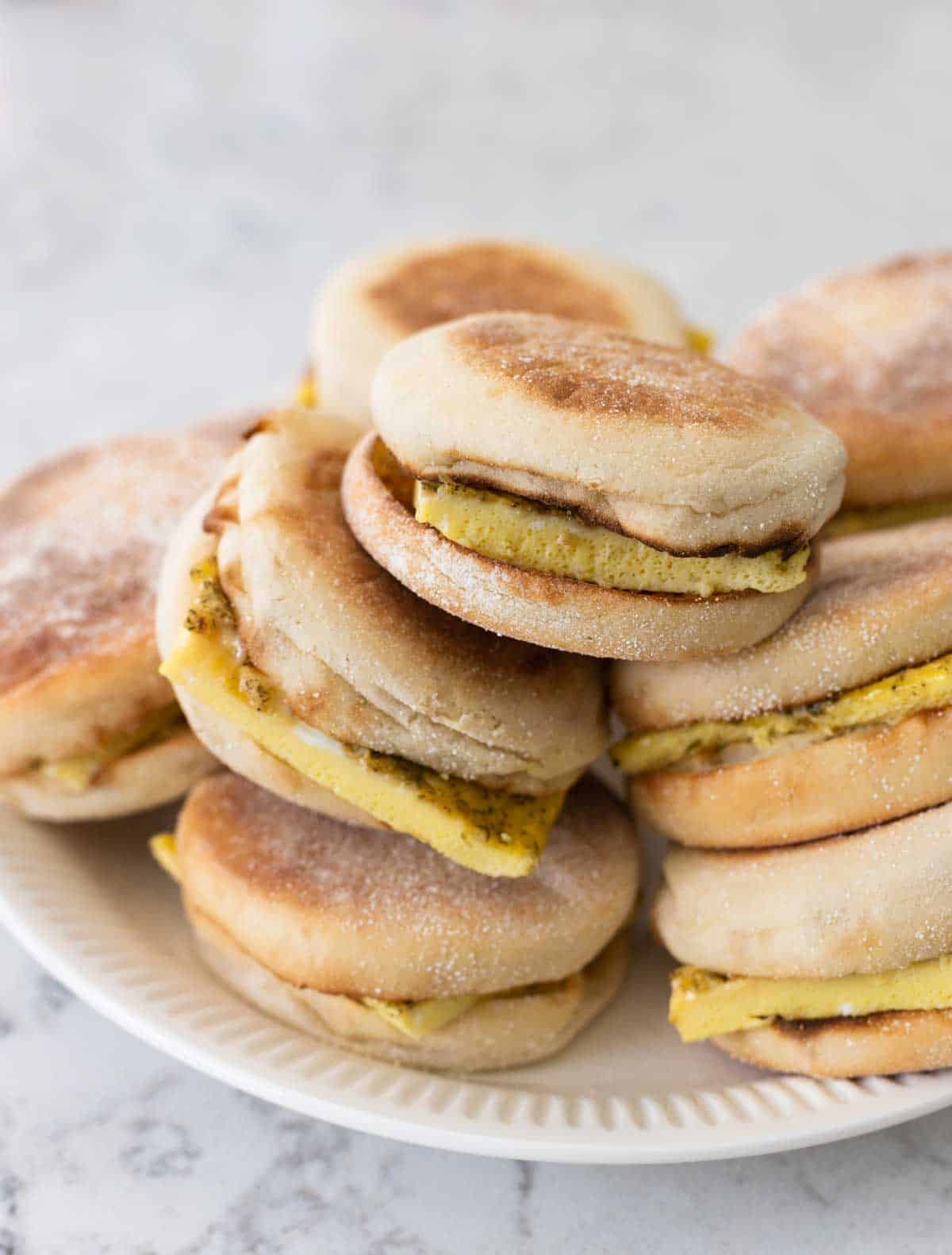 The plate has a pile of egg and english muffin sandwiches ready to be topped.