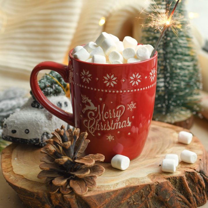 A red mug that says "Merry Christmas" has marshmallows on top and a sparkler. A pinecone rests in front.