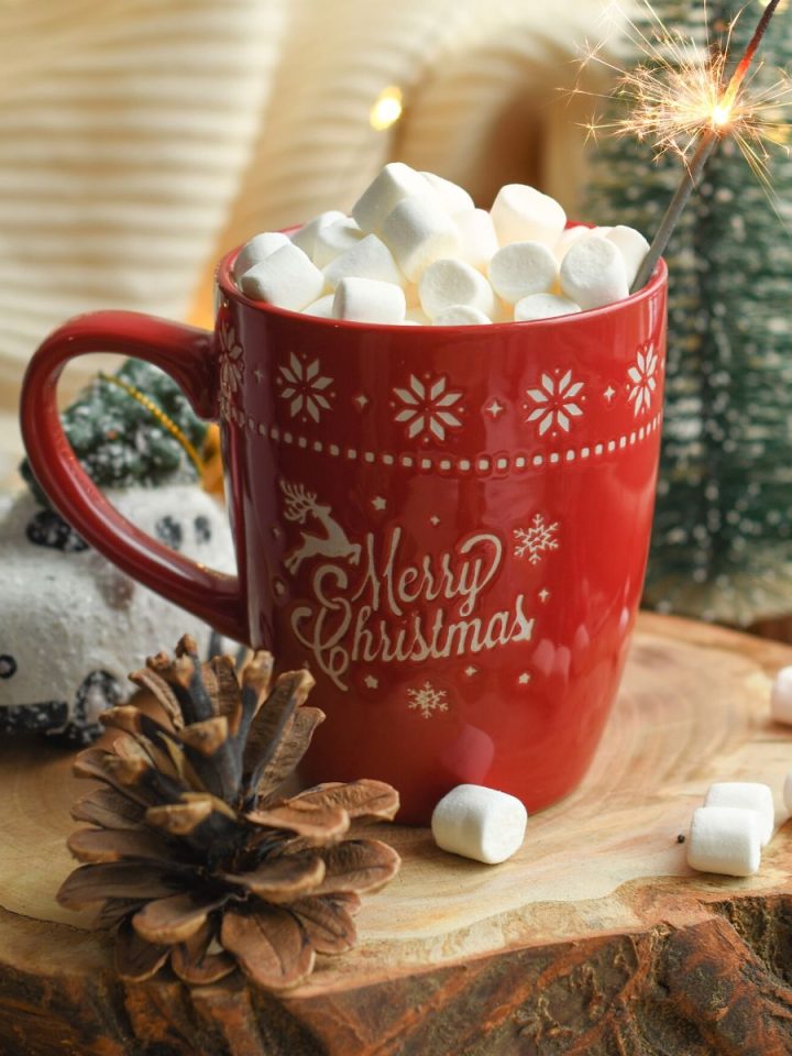 A red mug that says "Merry Christmas" has marshmallows on top and a sparkler. A pinecone rests in front.