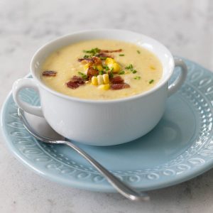 A white bowl of corn chowder sits on a blue plate. There is chunks of corn and bacon floating on top of the soup.