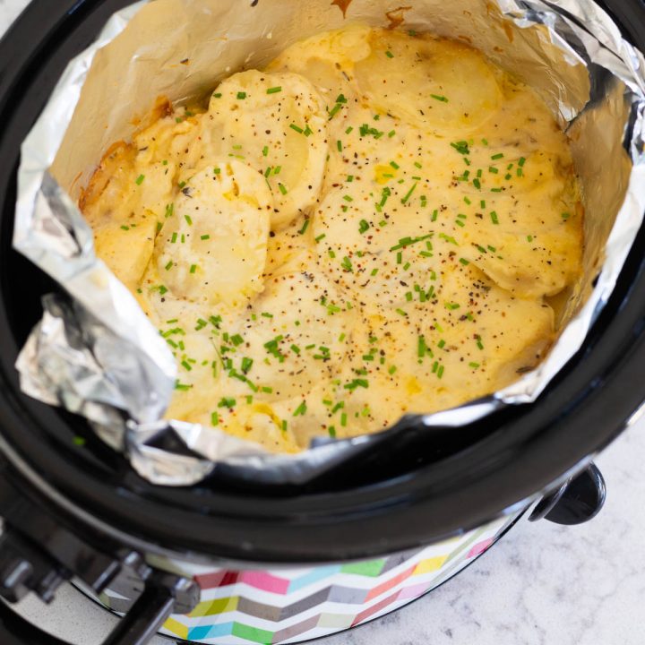 A slowcooker has an aluminum foil lining and is filled with baked scalloped potatoes with fresh chives sprinkled over the top.