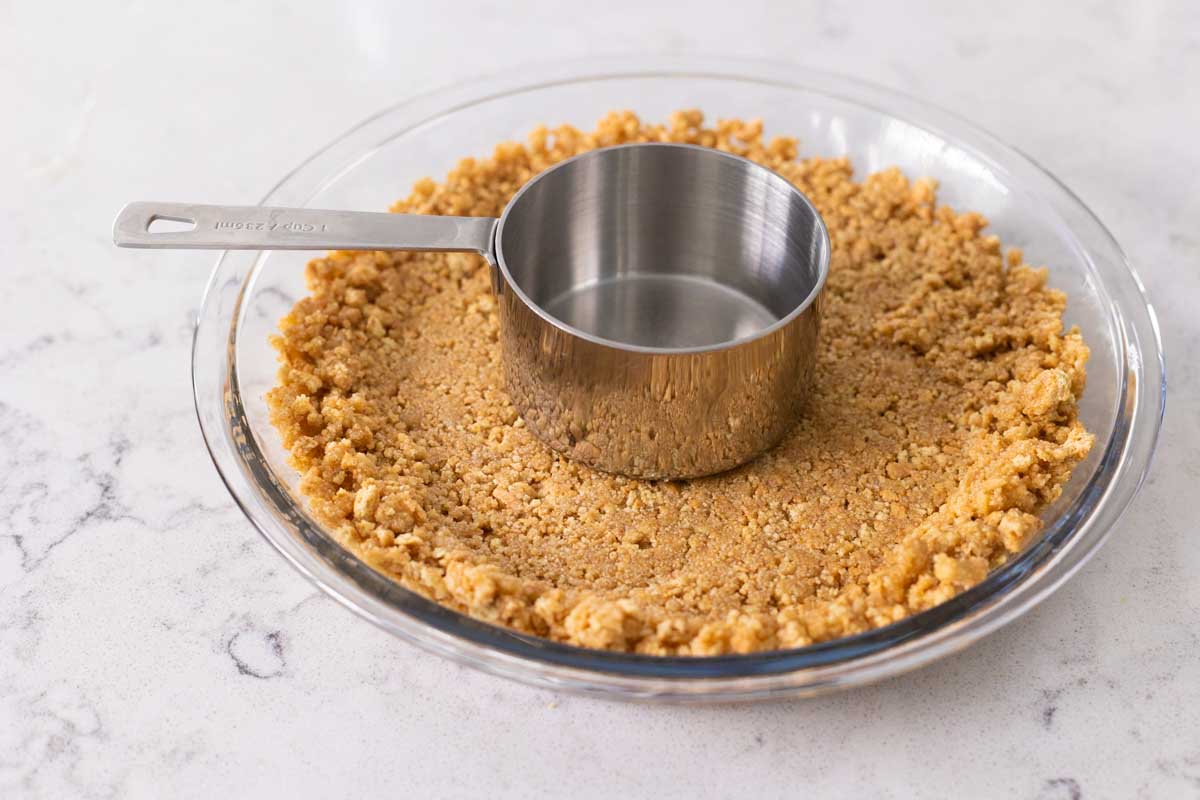 The pie plate is filled with the graham cracker crust and a measuring cup is being used to press it into place evenly.