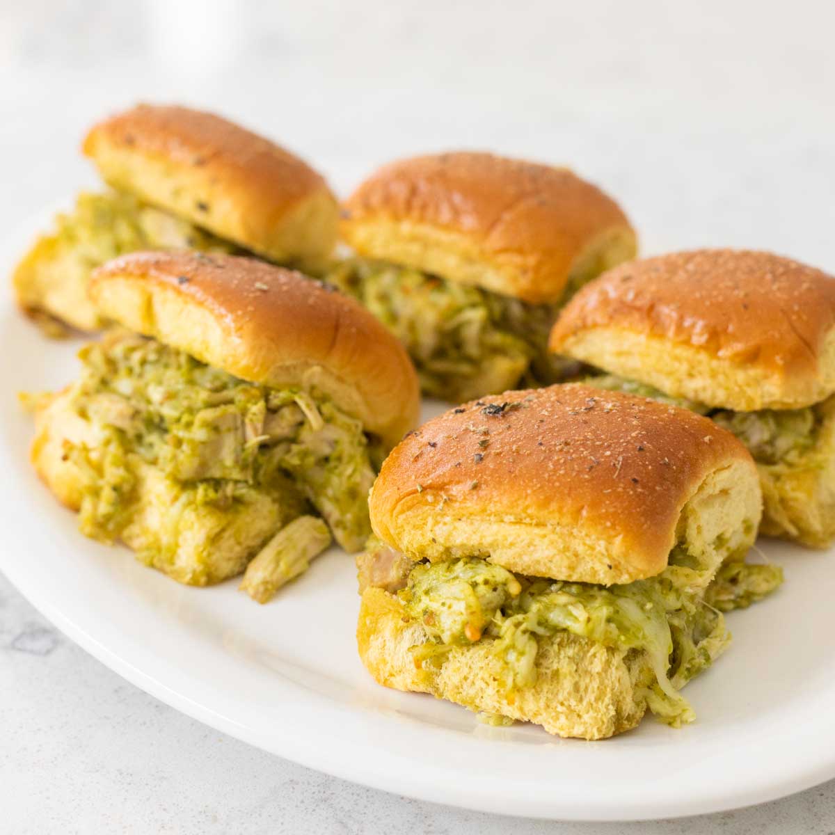 A white platter is filled with shredded chicken sliders with a pesto sauce.