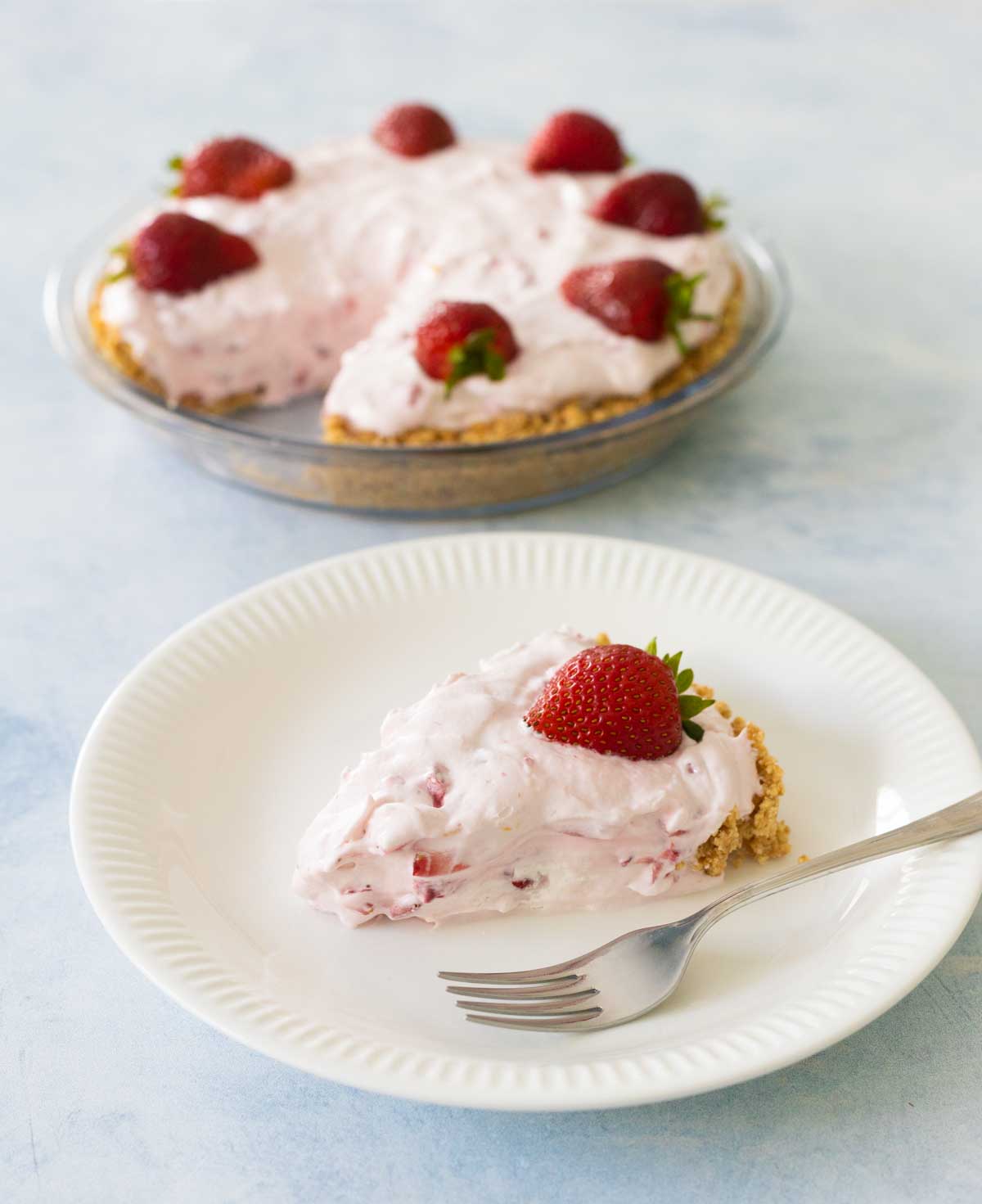 A slice of strawberry cream pie on a plate next to the pie in the background.