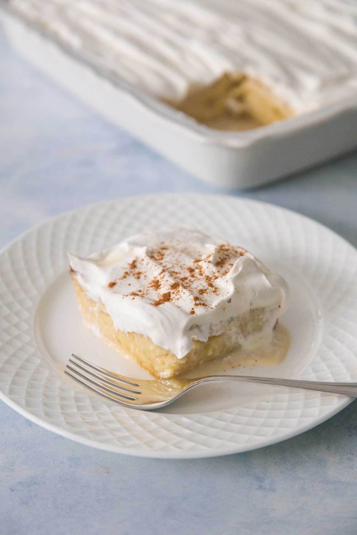 The serving of tres leches cake is on a plate, the cake pan is in the background and you can see the space where the cake slice is missing.