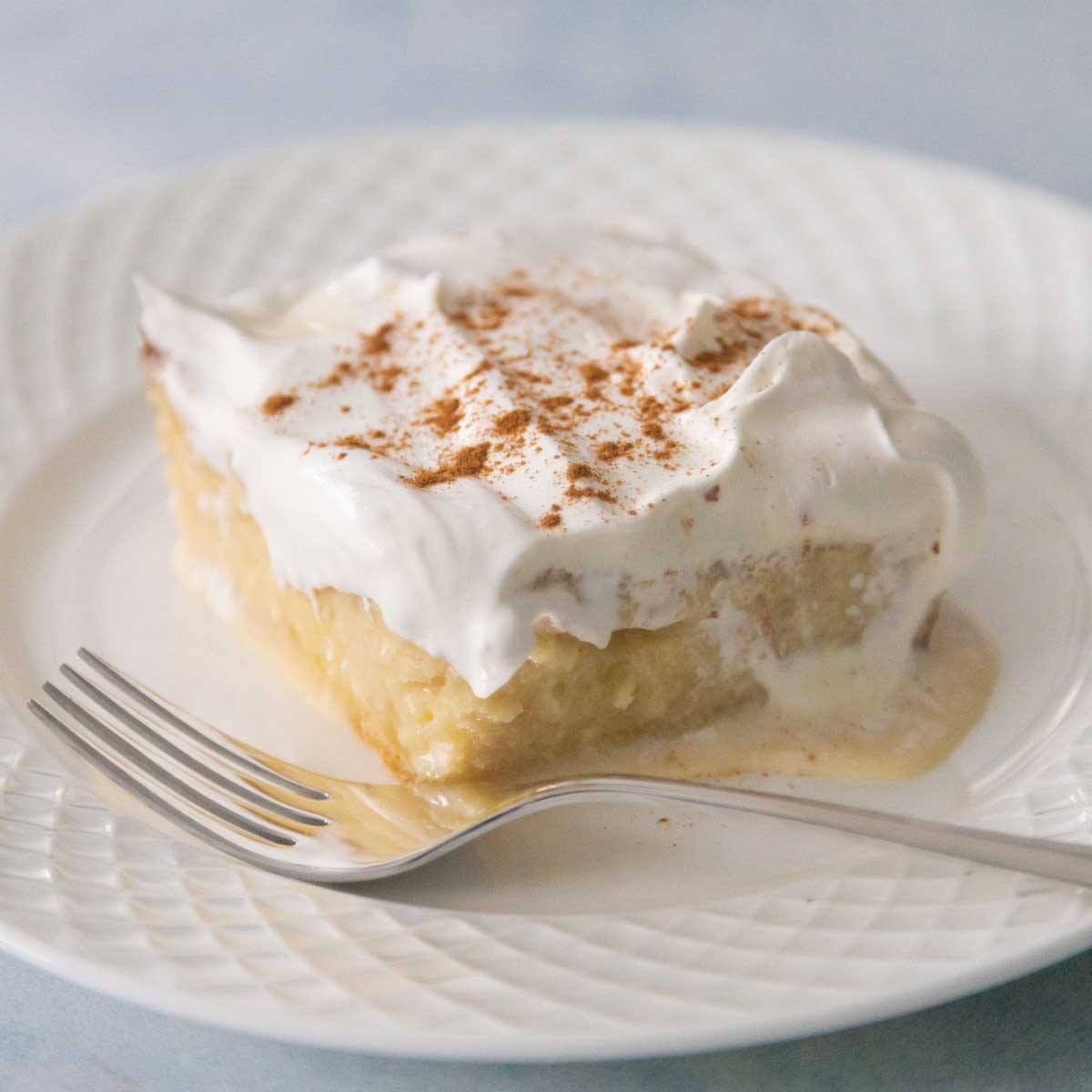 A square slice of tres leches cake is on a plate with a fork. You can see the three milks drizzling down the side of the vanilla cake and cinnamon is sprinkled on top of the whipped cream frosting.