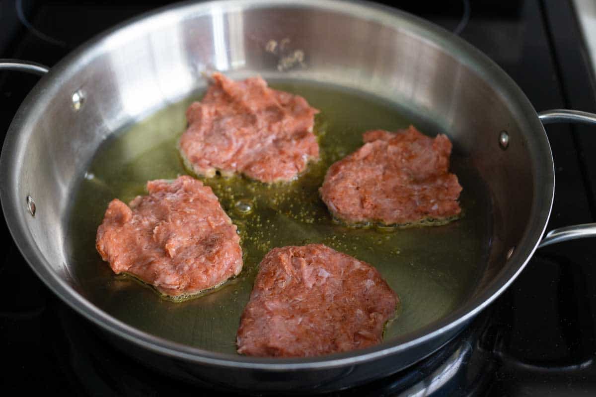 A large skillet has olive oil in it and 4 turkey breakfast sausage patties cooking on the first side.