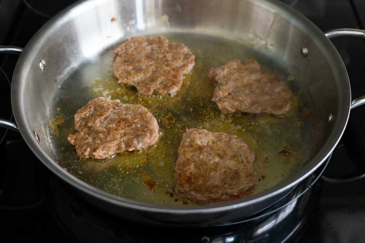 A large skillet has olive oil in it and 4 turkey breakfast sausage patties cooking on the second side.