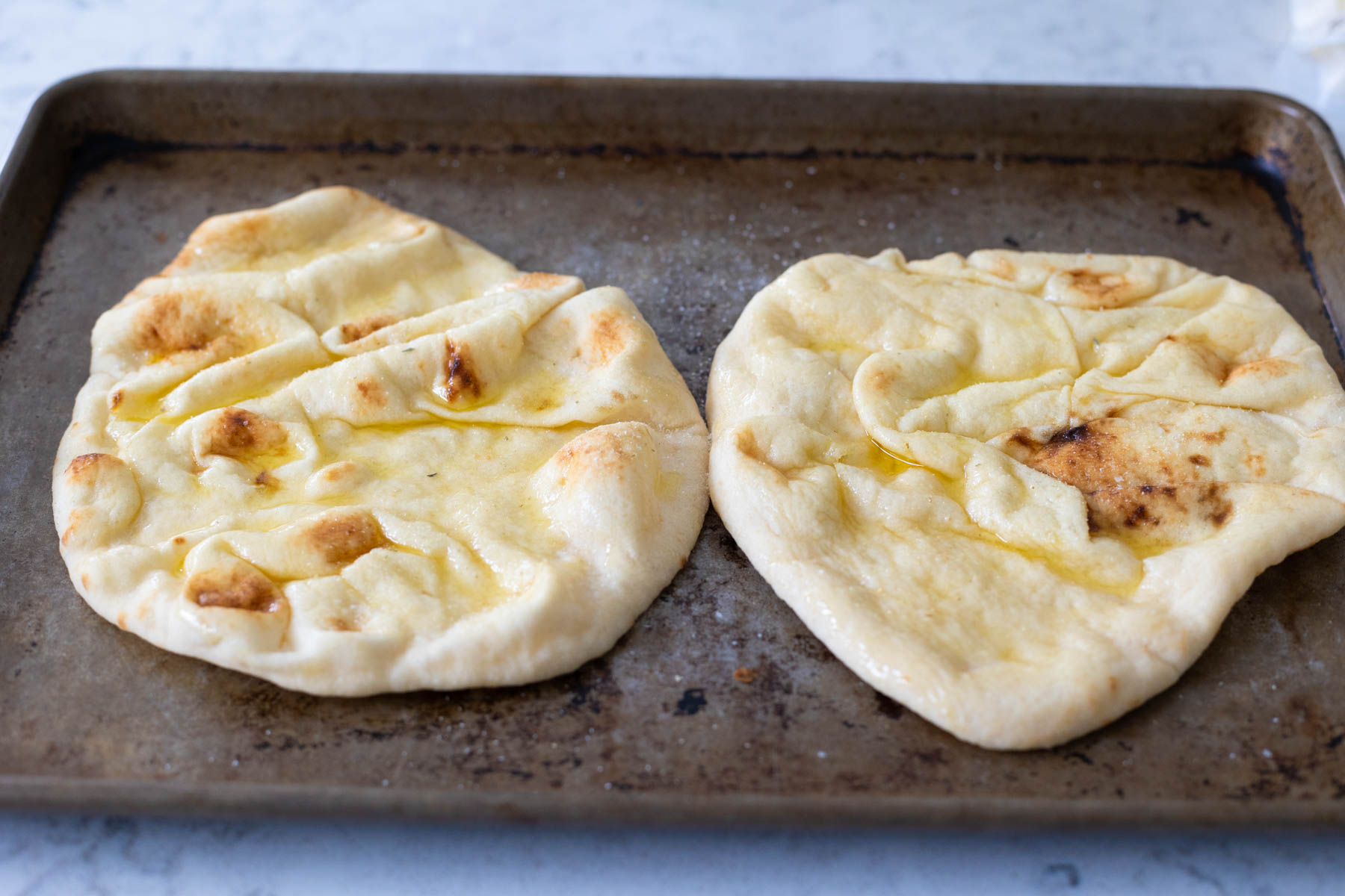 Two naan breads are on a large baking sheet and have been brushed with olive oil.