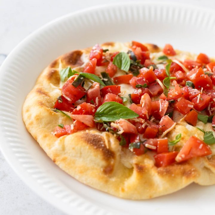 The bruschetta pizza is served on a white plate. Fresh tomatoes and basil are scattered over the top.