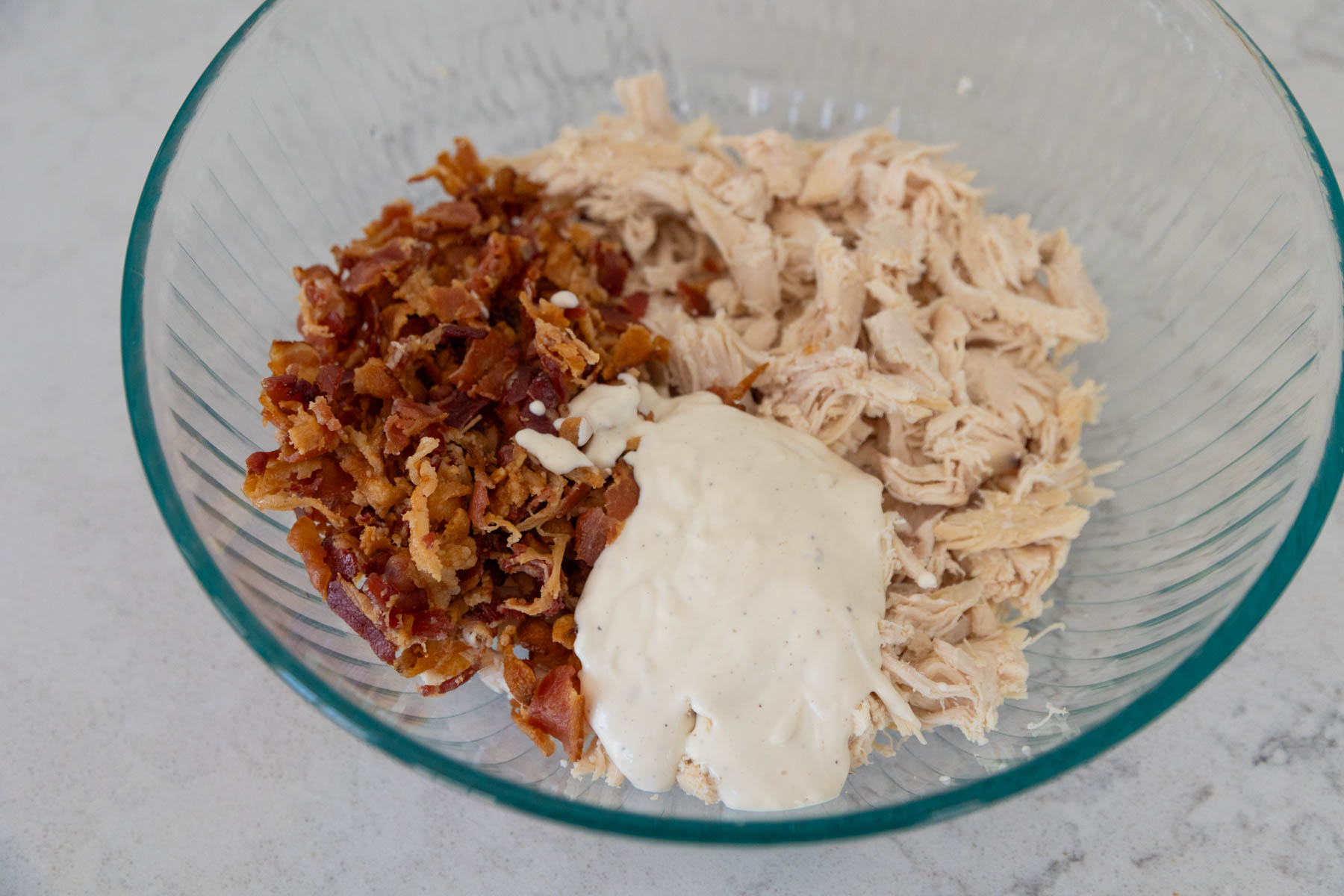 The shredded rotisserie chicken meat is in a mixing bowl with crumbled bacon and ranch dressing.
