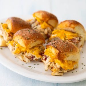 A white platter has several of the chicken bacon ranch sliders with melted cheese and golden brown tops.