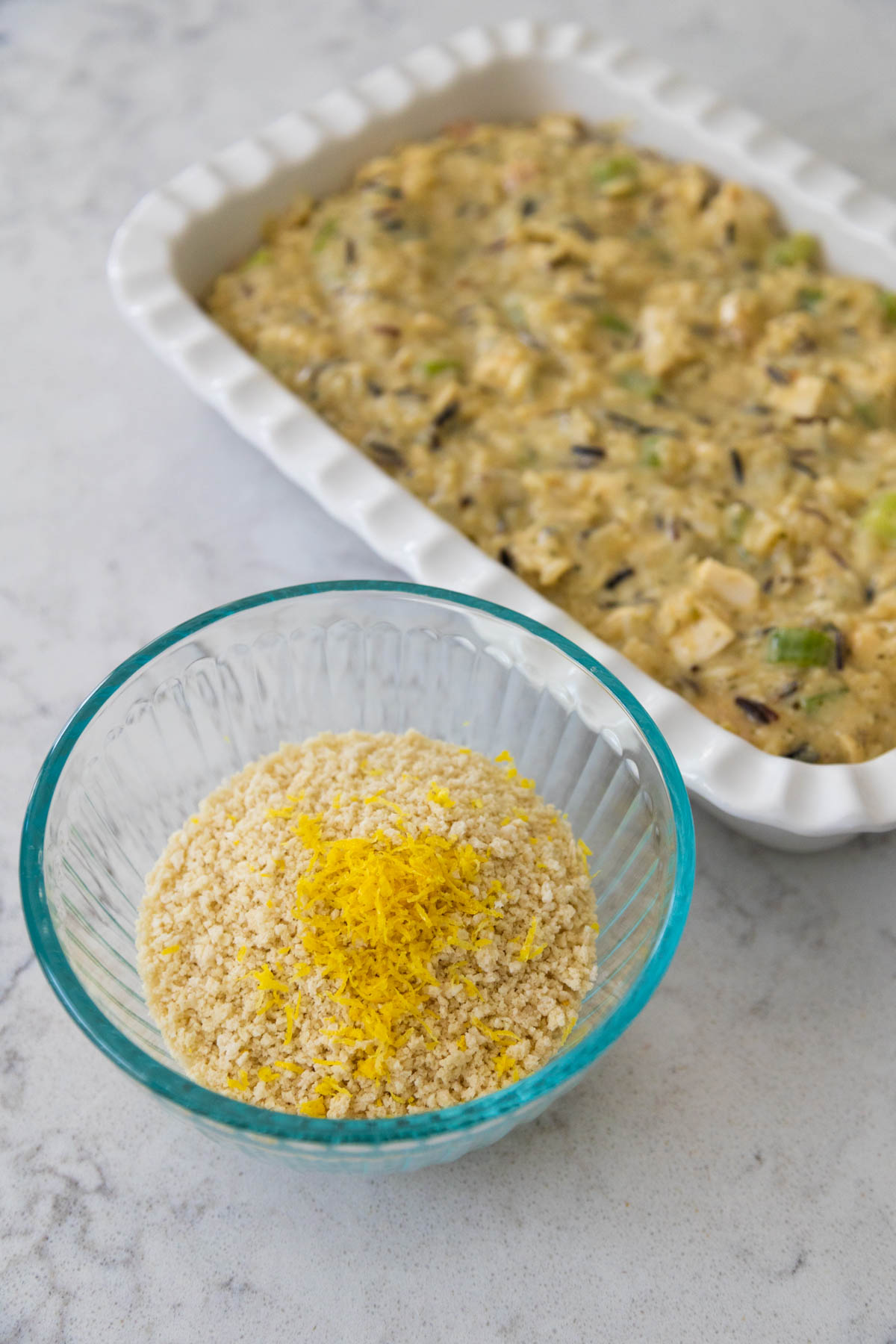 A bowl of breadcrumbs with lemon zest on top sits next to the baking dish filled with creamy chicken.