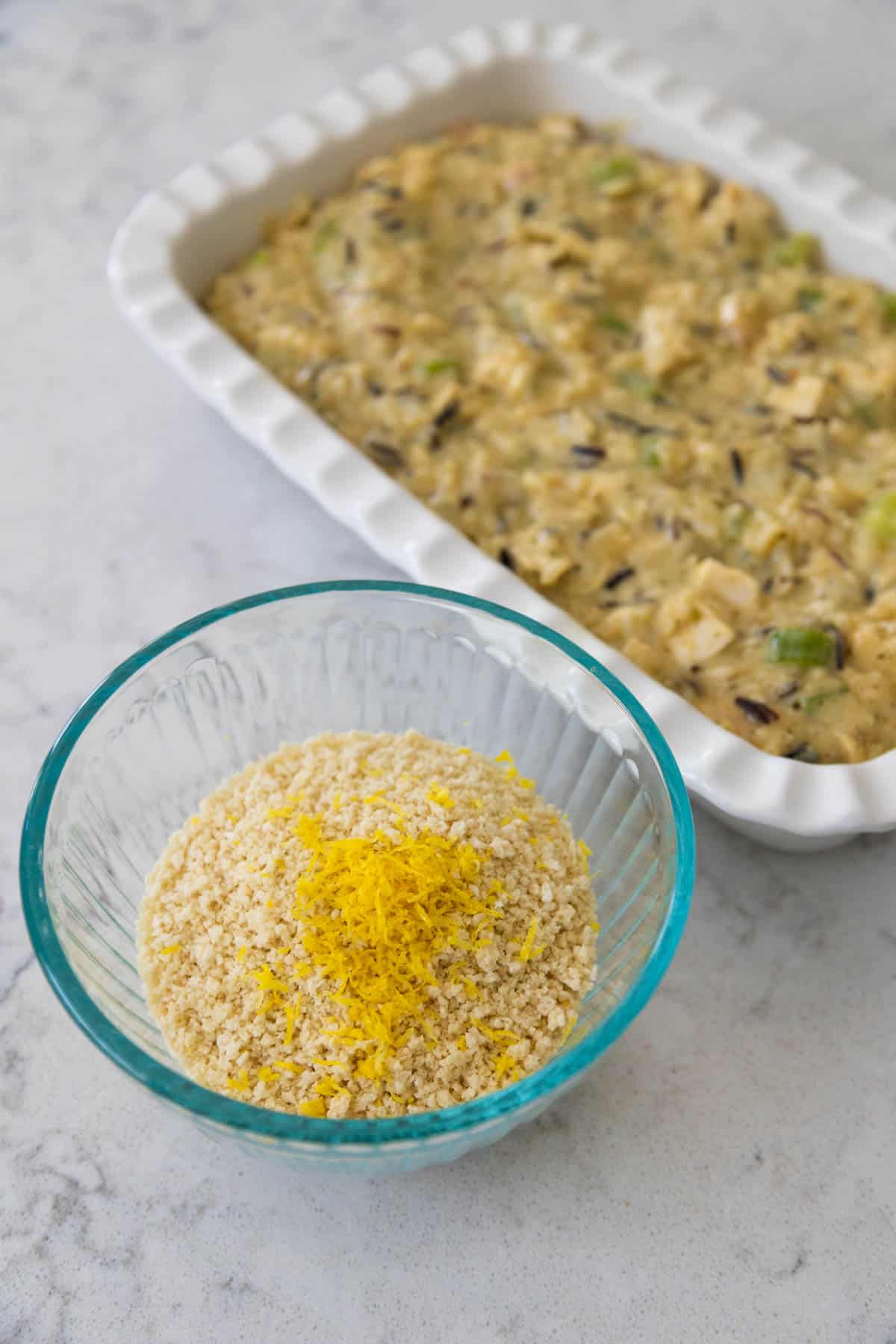 A bowl of breadcrumbs with lemon zest on top sits next to the baking dish filled with creamy chicken.