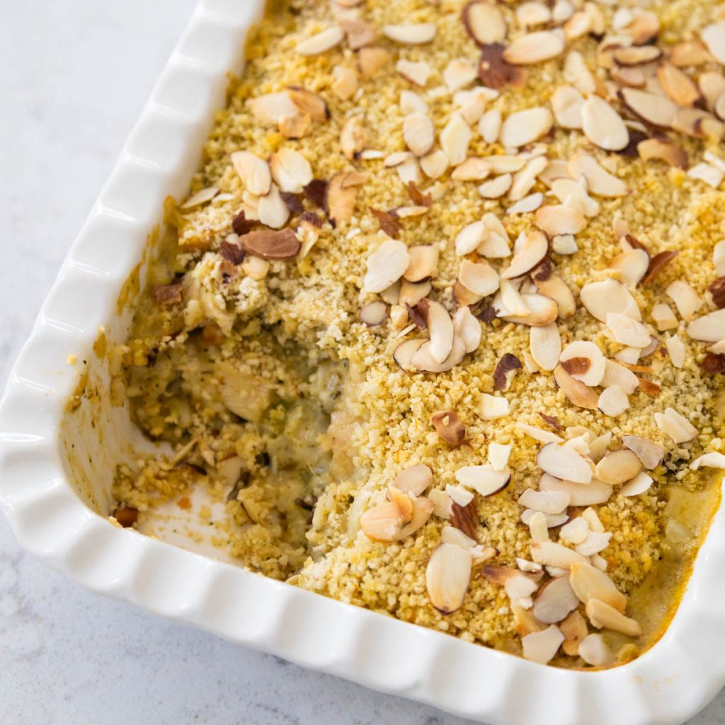 A white baking dish has chicken wild rice casserole topped with bread crumbs and almonds. A scoop is missing.
