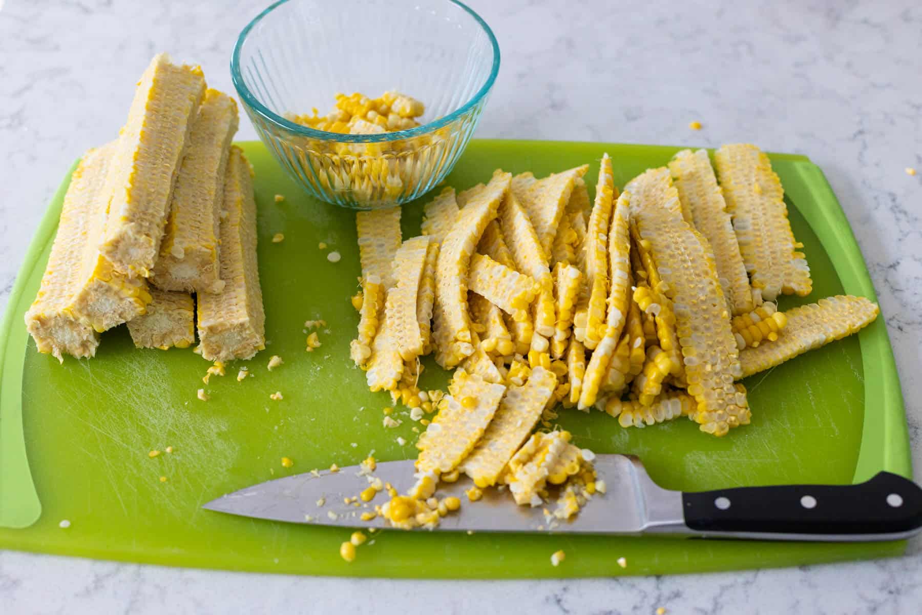 The corn has been removed from the cobs by a large chef knife. The kernels sit on the cutting board to the right, the empty cobs on the left.