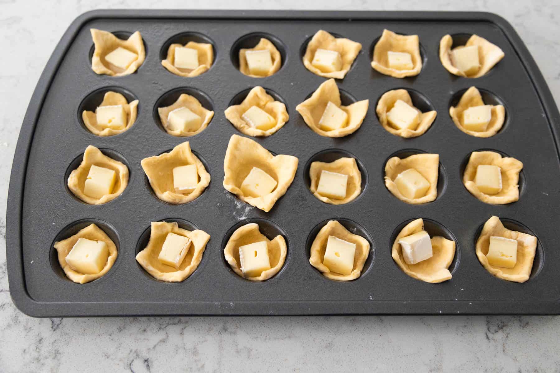 The squares of dough have been tucked into a mini muffin pan and brie cheese cubes added to each well.