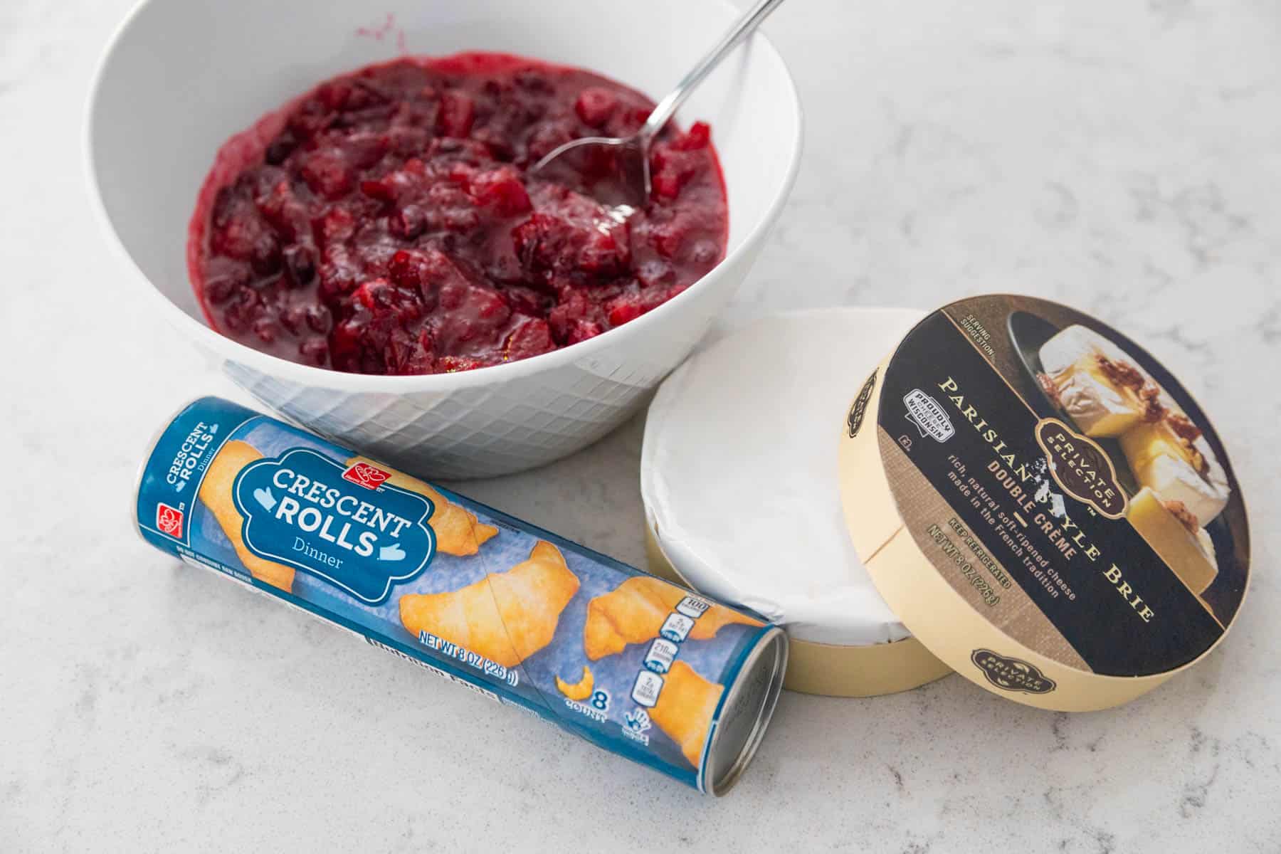 A can of crescent roll dough, a wheel of brie cheese, and a bowl of cranberry sauce.