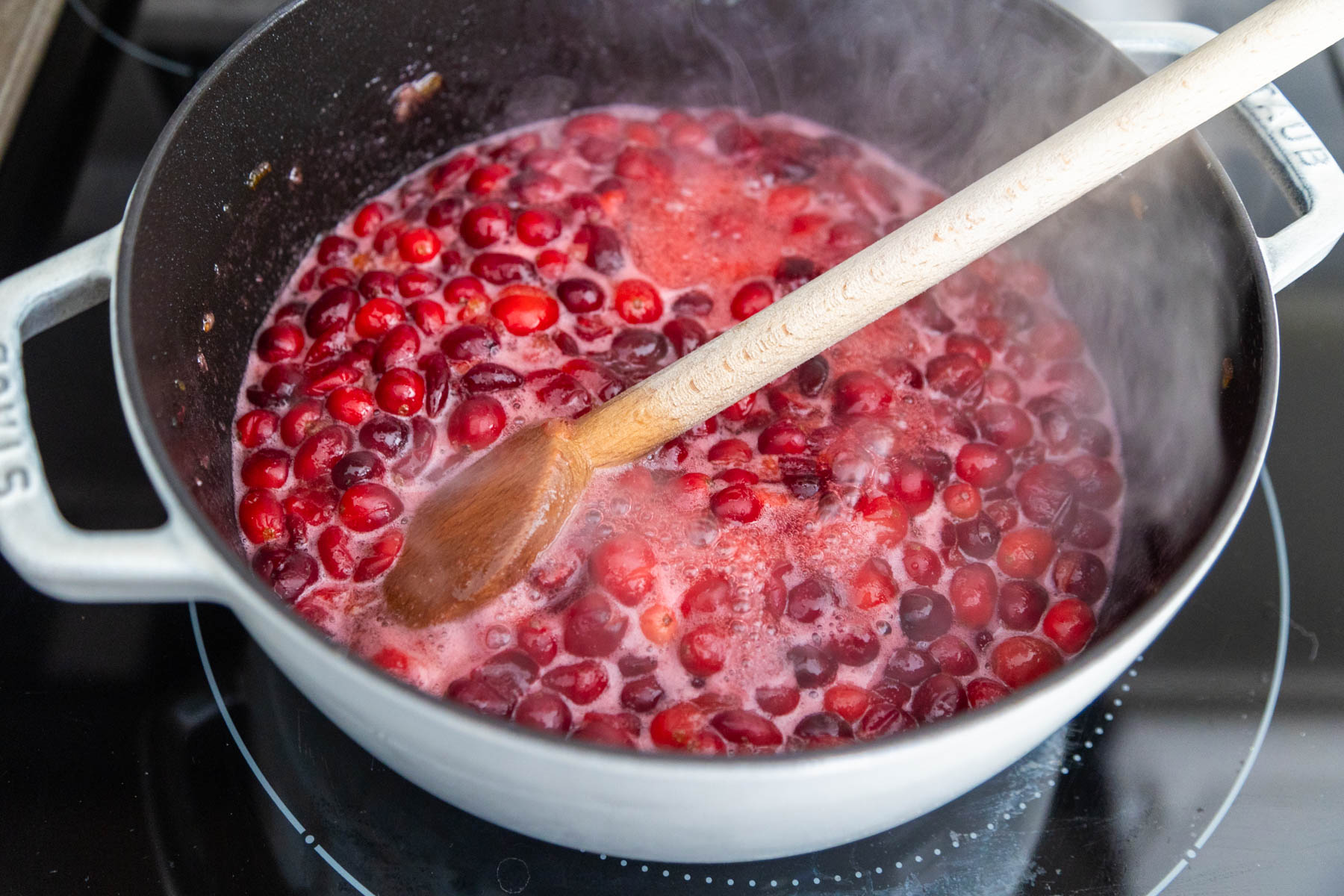 The cranberries are in a sauce pan bubbling. A wooden spoon is stirring them. 