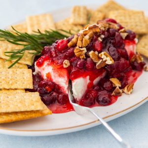 A platter has a brick of cream cheese with cranberry sauce spooned over the top. Chopped pecans are used for garnish and crackers and fresh rosemary circle the dip.
