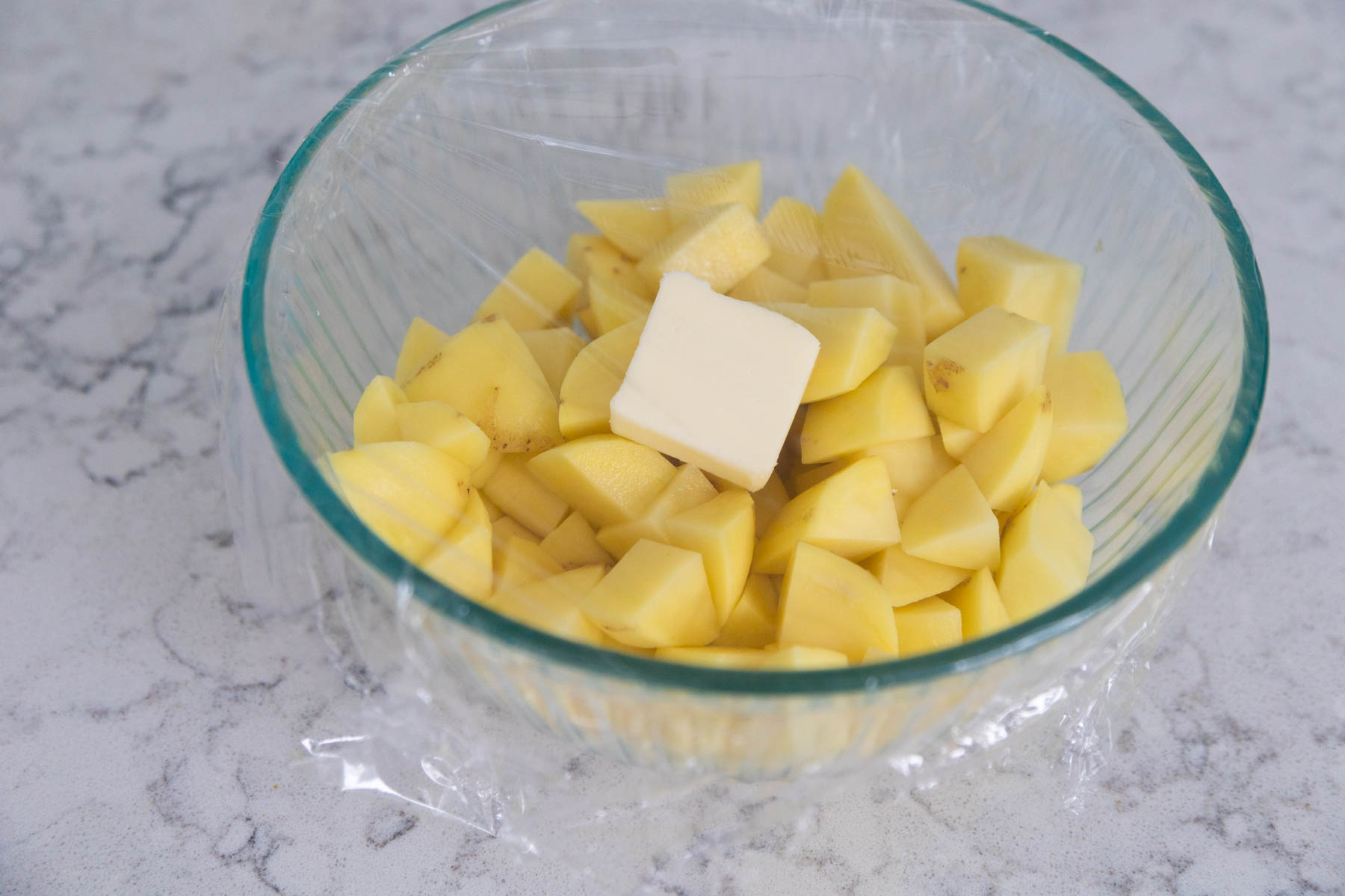 A mixing bowl with potato chunks and a pat of butter are covered with plastic wrap. 