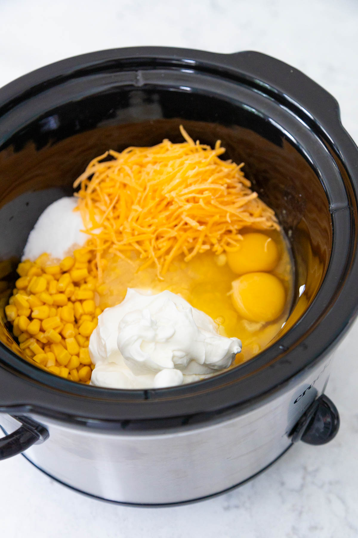 The corn, sour cream, eggs, shredded cheese, and sugar are in the bowl of the Crock Pot.