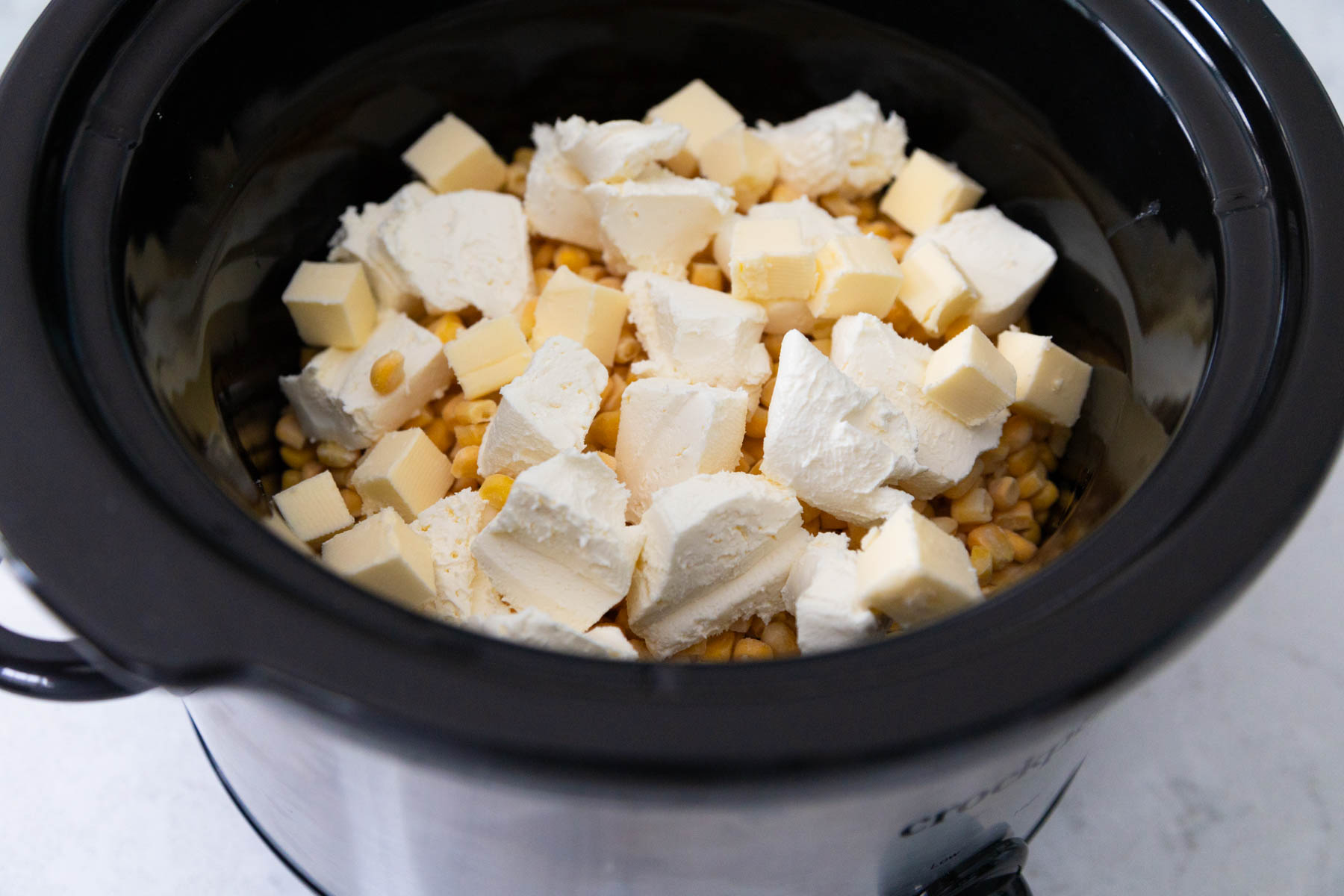 The crock pot bowl is filled with frozen corn and topped with cubes of cream cheese and butter.