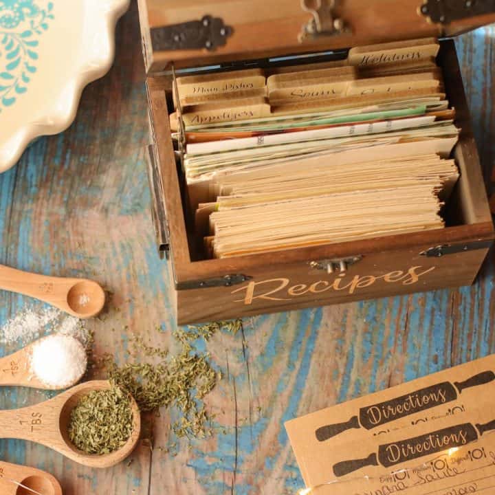 An old fashioned woodend recipe box is filled with recipe cards. Measuring spoons filled with salt and herbs are scattered around it.