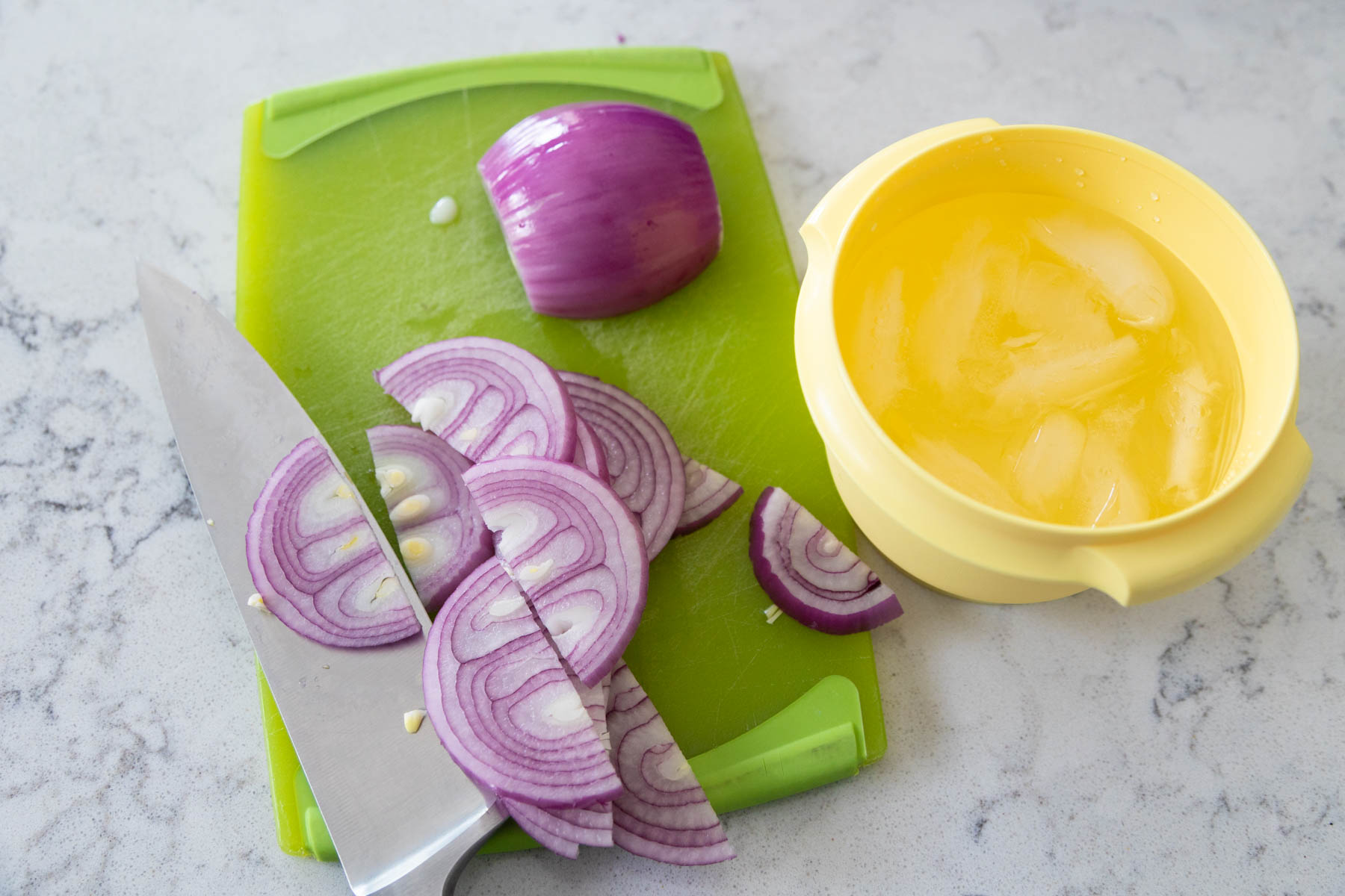 The red onion has been sliced on a cutting board and a yellow bowl of ice water is ready for it to do a quick soak.