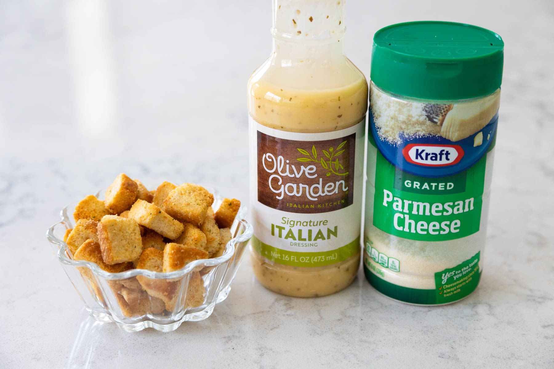 A bottle of Olive Garden Italian dressing sits next to a bowl of croutons and a jar of parmesan cheese.