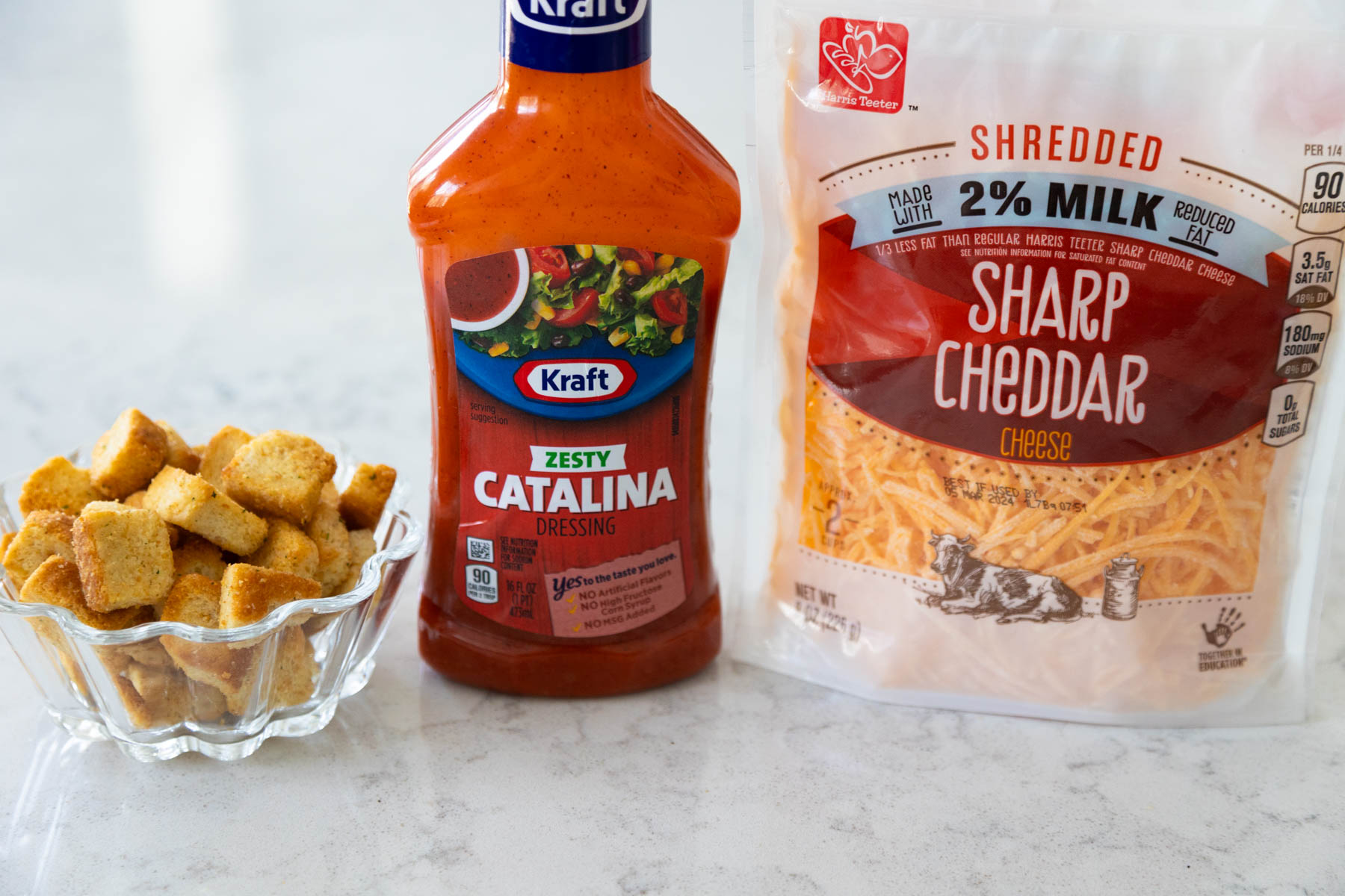 A bottle of Catalina dressing sits next to a bag of shredded cheddar cheese and a bowl of croutons.