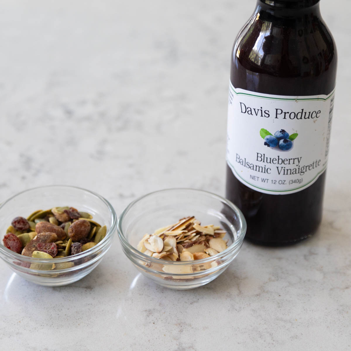 A bottle of blueberry vinaigrette sits next to a bowl of toasted almonds and a bowl of seasoned almonds with dried cranberries.