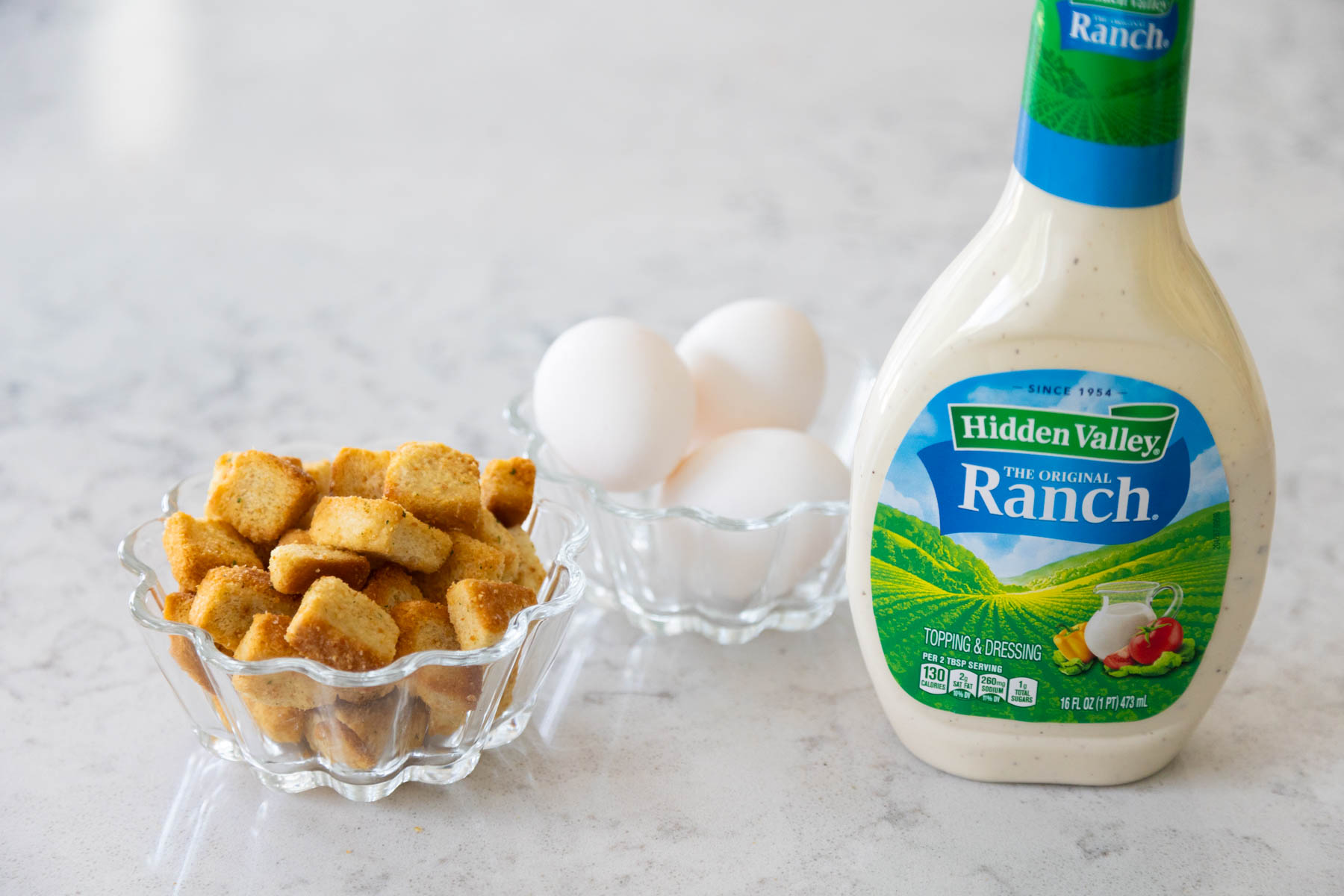A bottle of ranch dressing sits next to a bowl of hardboiled eggs and a bowl of croutons.