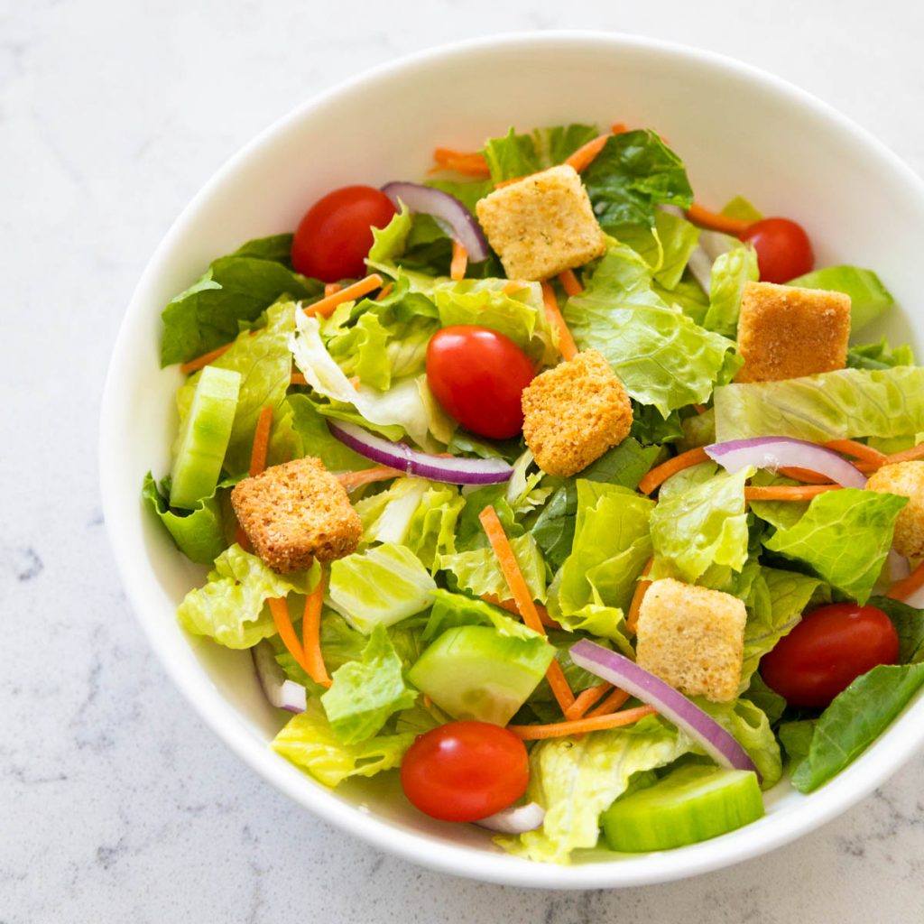 The fresh garden salad is served in a white bowl. You can see lettuce, cherry tomatoes, croutons, and cucumber.