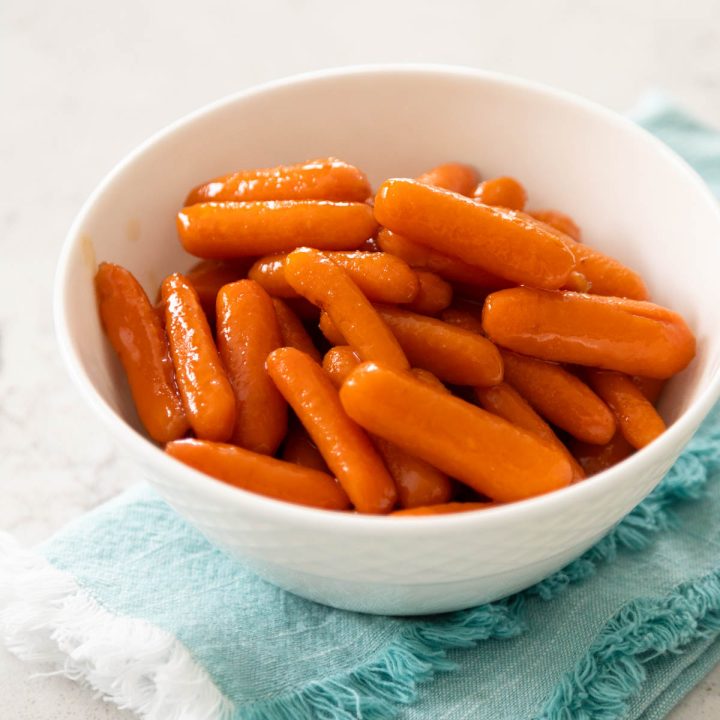 A white bowl is filled with glazed baby carrots, a blue napkin is tucked underneath.