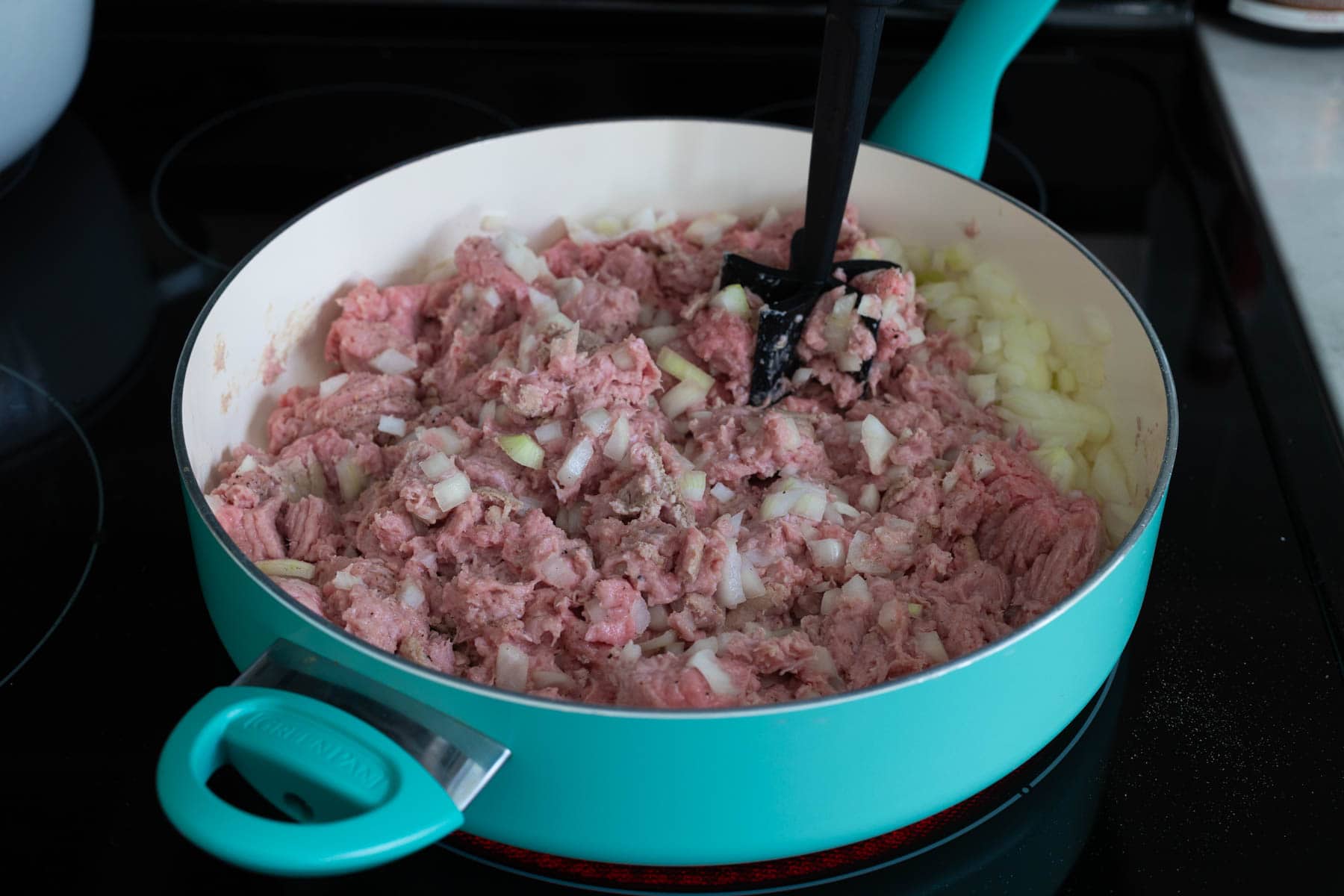 The ground turkey and chopped onions are in a large skillet being chopped by a hand tool.