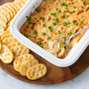 A white baking dish with golden brown hot crab dip has a spoon serving up a portion. There are butter crackers all around the dish.