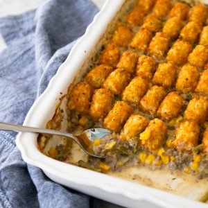 The tater tot casserole has a spoon scooping a serving, you can see the ground beef, green beans, and corn with golden brown tater tots on top.
