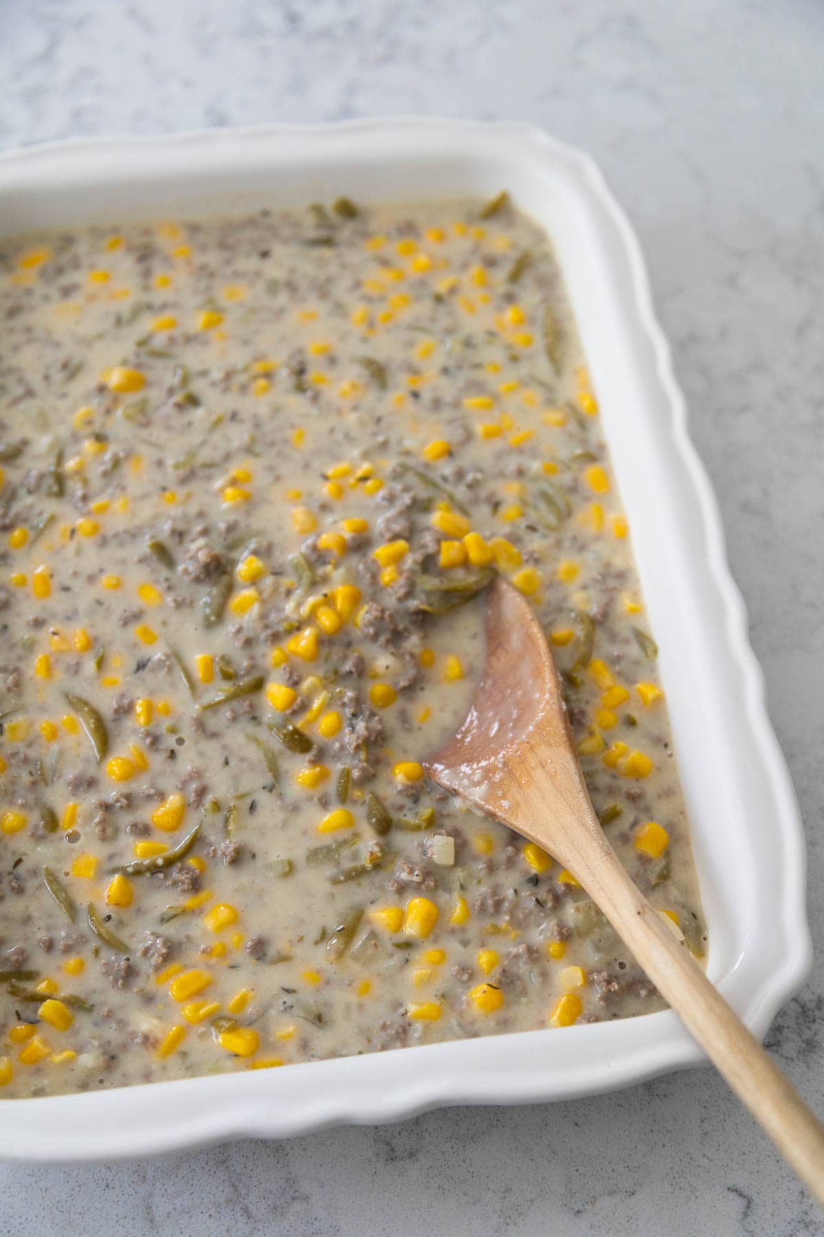 The ground beef and vegetables base have been spread into a white baking dish with a wooden spoon.