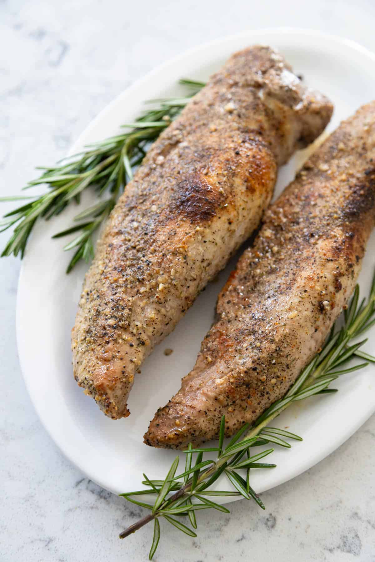 The pork tenderloins are roasted and now served on a white platter with fresh rosemary for garnish.