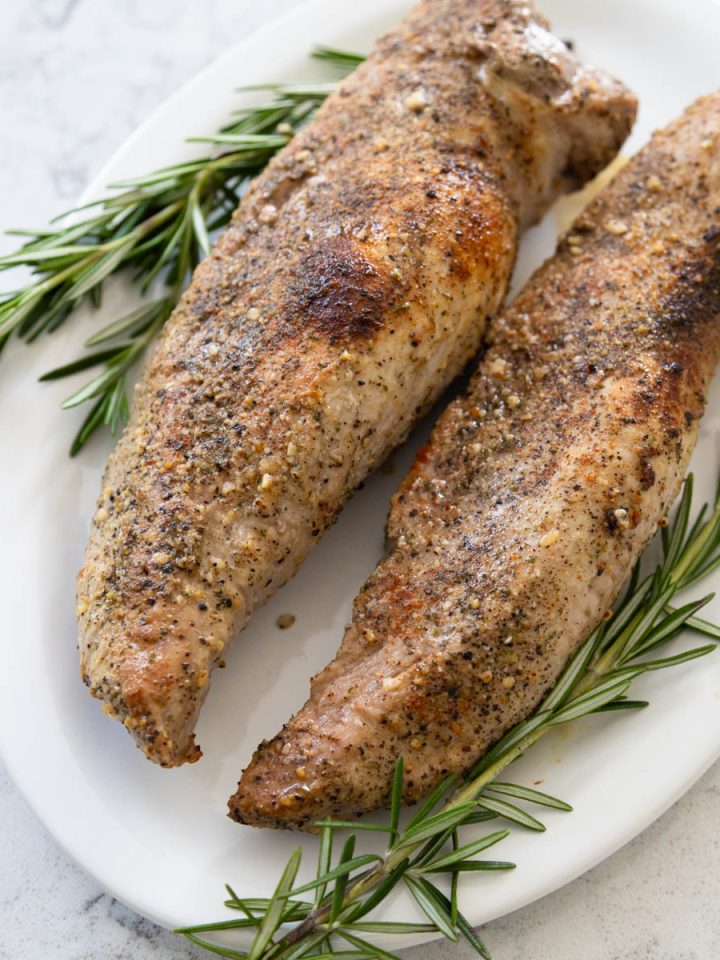 Two Smithfield pork tenderloins have been seasoned and roasted. They are now served on a white platter with fresh rosemary garnish.