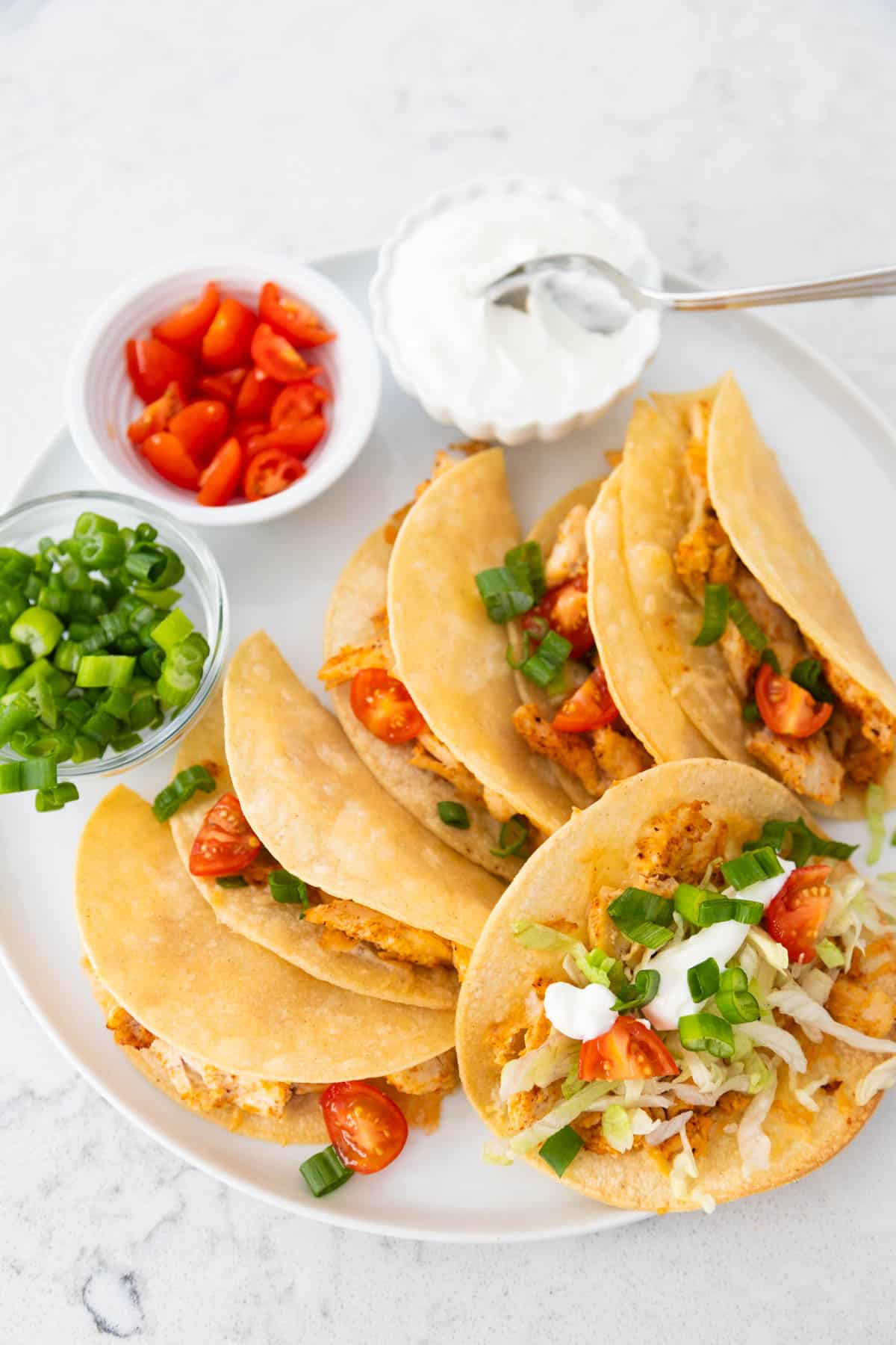 The platter of tacos has fresh tomatoes, green onions, and sour cream in little bowls.