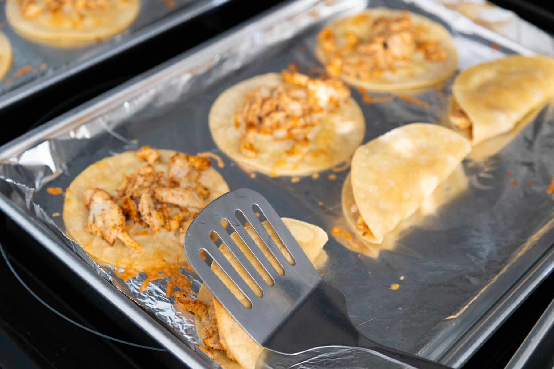 A spatula is folding the tacos into shape on the baking sheet.