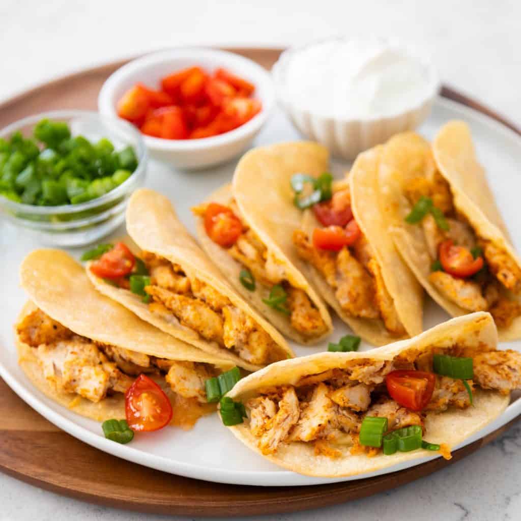 A platter of crispy baked chicken tacos has bowls of tomatoes, green onions, and sour cream in the background for garnishing.