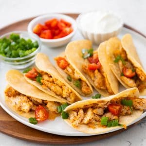 A platter of crispy baked chicken tacos has bowls of tomatoes, green onions, and sour cream in the background for garnishing.