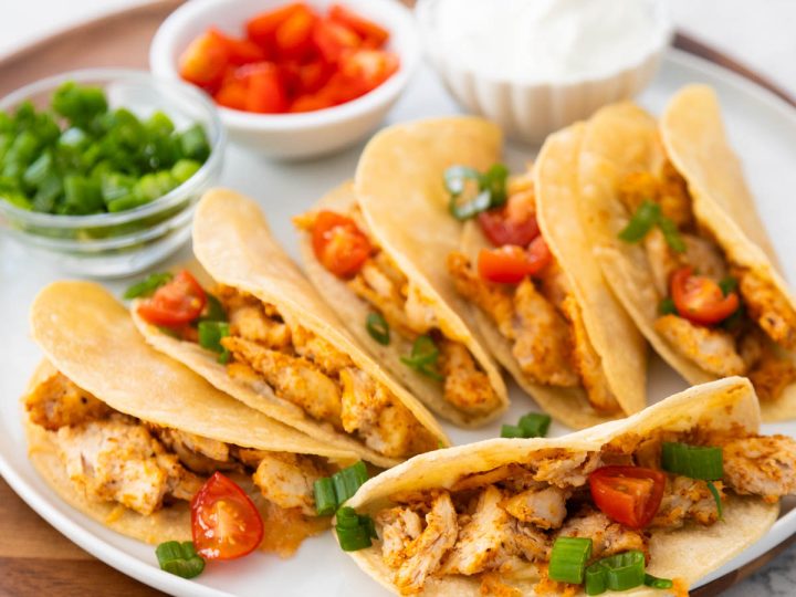 A platter of crispy baked chicken tacos has bowls of tomatoes, green onions, and sour cream in the background for garnishing.