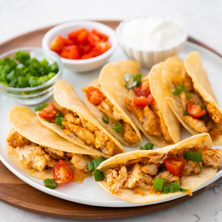 A platter of crispy baked chicken tacos has bowls of tomatoes, green onions, and sour cream in the background for garnishing.