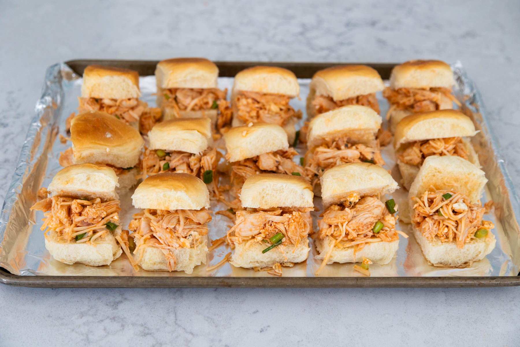 The assembled chicken sliders are on a large baking pan lined with aluminum foil.